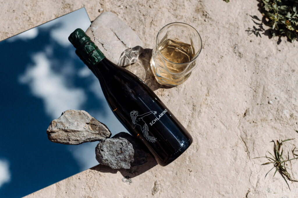Schlawino-Flasche und ein Glas Weißwein auf Sand, daneben Spiegel und Muschel